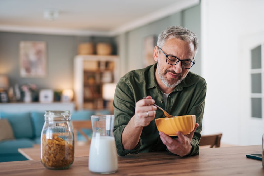 An elderly man eats cornflakes with milk for breakfast, sitting in the modern apartment